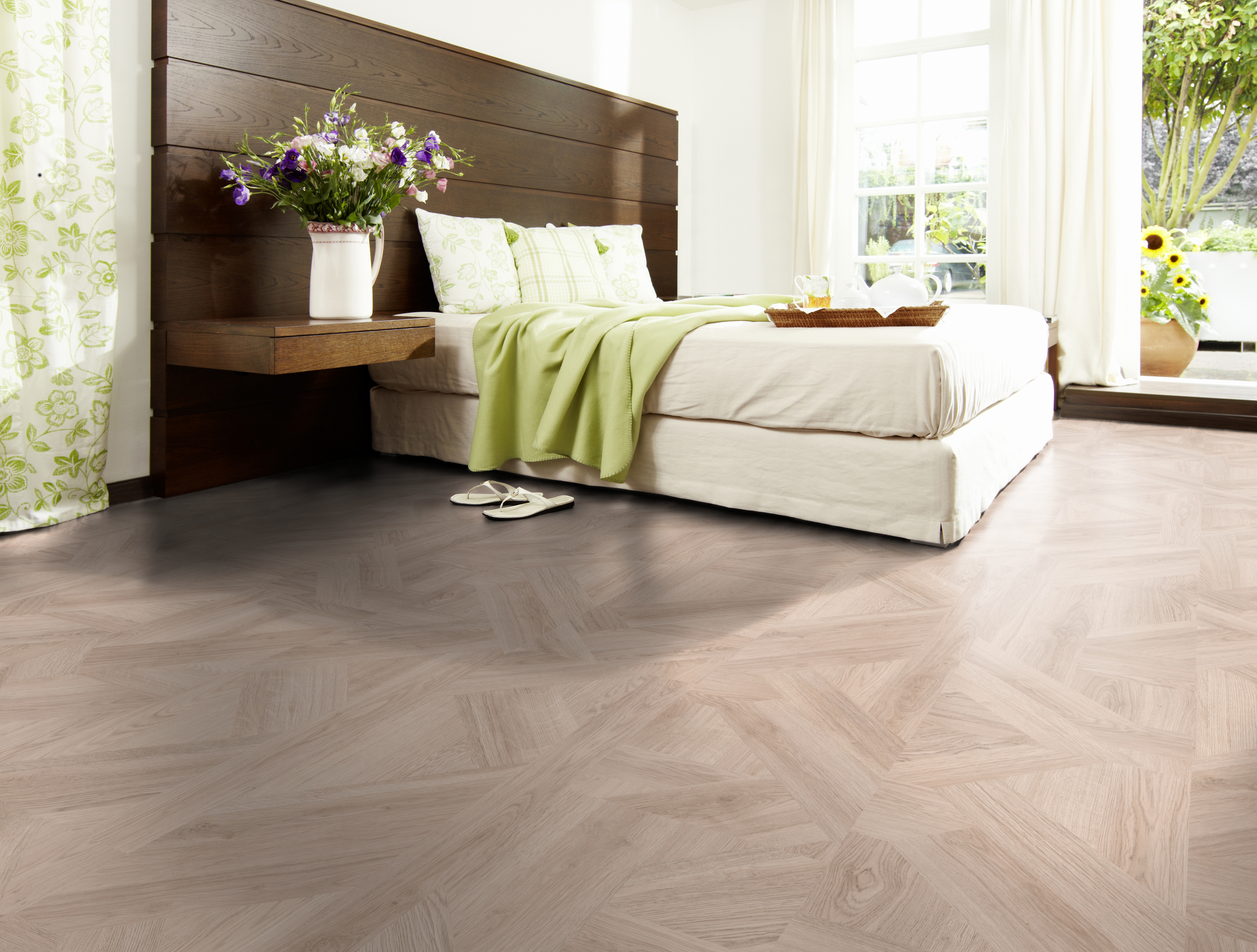 Schlafzimmer mit Bett, Holz-Kopfteil und Bodenbelag in geometrischer Holzoptik, Wandgestaltung mit hellen Vorhängen.